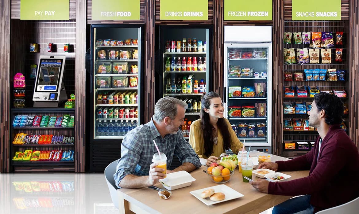 Three people dining in a modern breakroom with stocked food, drink, and snack displays.